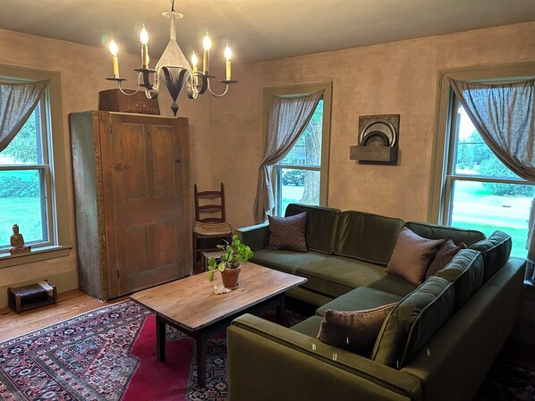 A cozy living room with a wooden cabinet, a green sofa, a small table with a plant, and a chandelier. - Charming 3-bedroom farmhouse in welcoming Skaneateles, short walk to village. (Skaneateles)