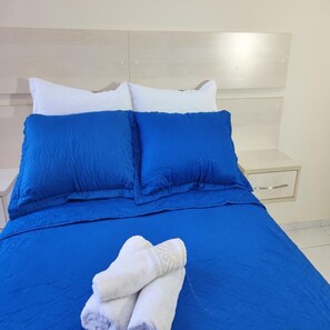 Een bed met blauw beddengoed en witte kussens, een witte nachtkastje met een lade, en een opgevouwen witte handdoek op het bed.