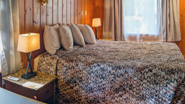 Ein Schlafzimmer einer Holzhütte mit einem Bett, zwei Nachttischlampen, einem Nachttisch und einem Fenster mit Vorhängen.