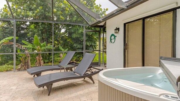 A covered outdoor area with two lounge chairs, a hot tub, and a glass enclosure with a view of lush greenery. - Pool - 3 bedrooms - 2 baths - Pet Friendly - 3 Miles to Beach (Bonita Springs)