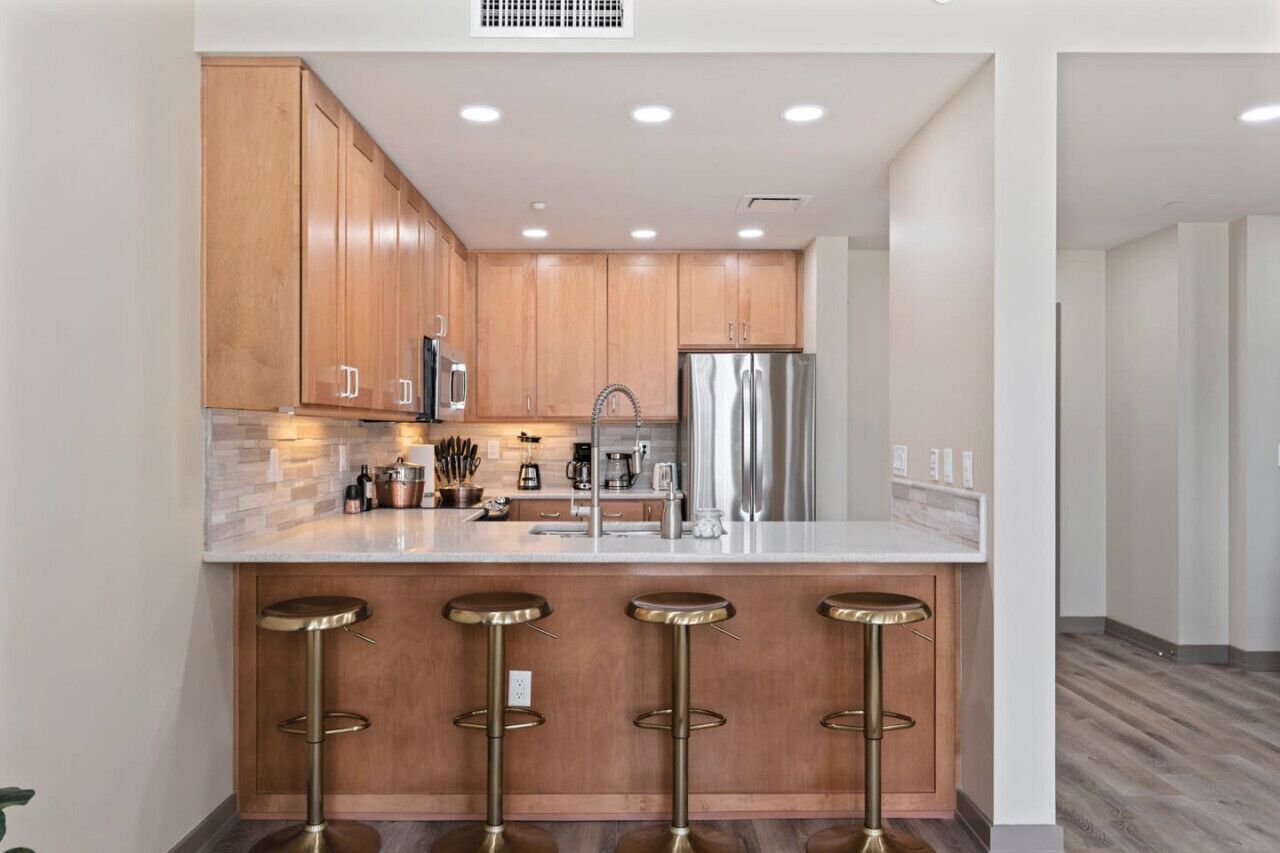 A modern kitchen with wooden cabinets, stainless steel appliances, a marble countertop, and three bar stools.