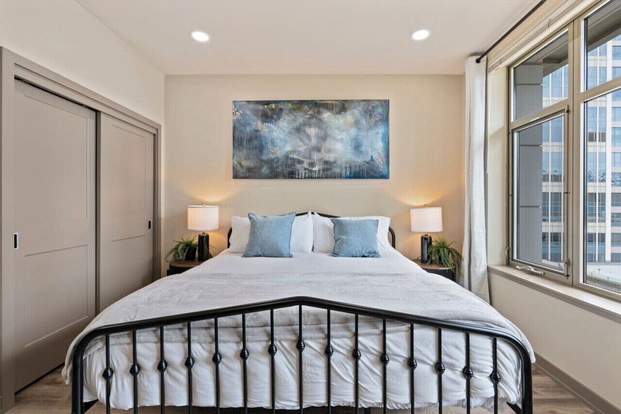 A bedroom with a large bed, two bedside tables with lamps, a painting on the wall, and a window with a view of buildings.