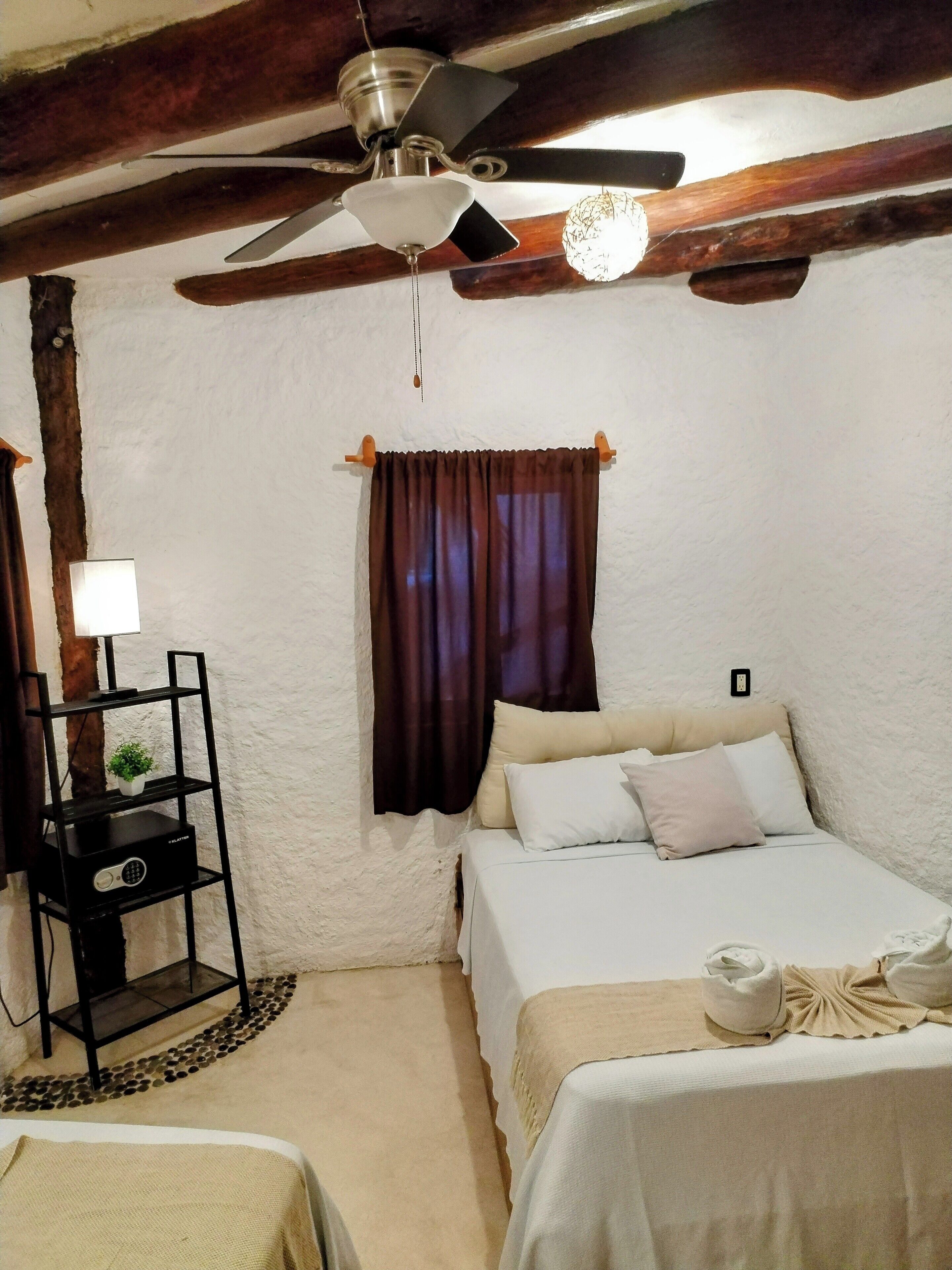A bedroom with a bed, a ceiling fan, a small table with a lamp, and a shelf with a plant.