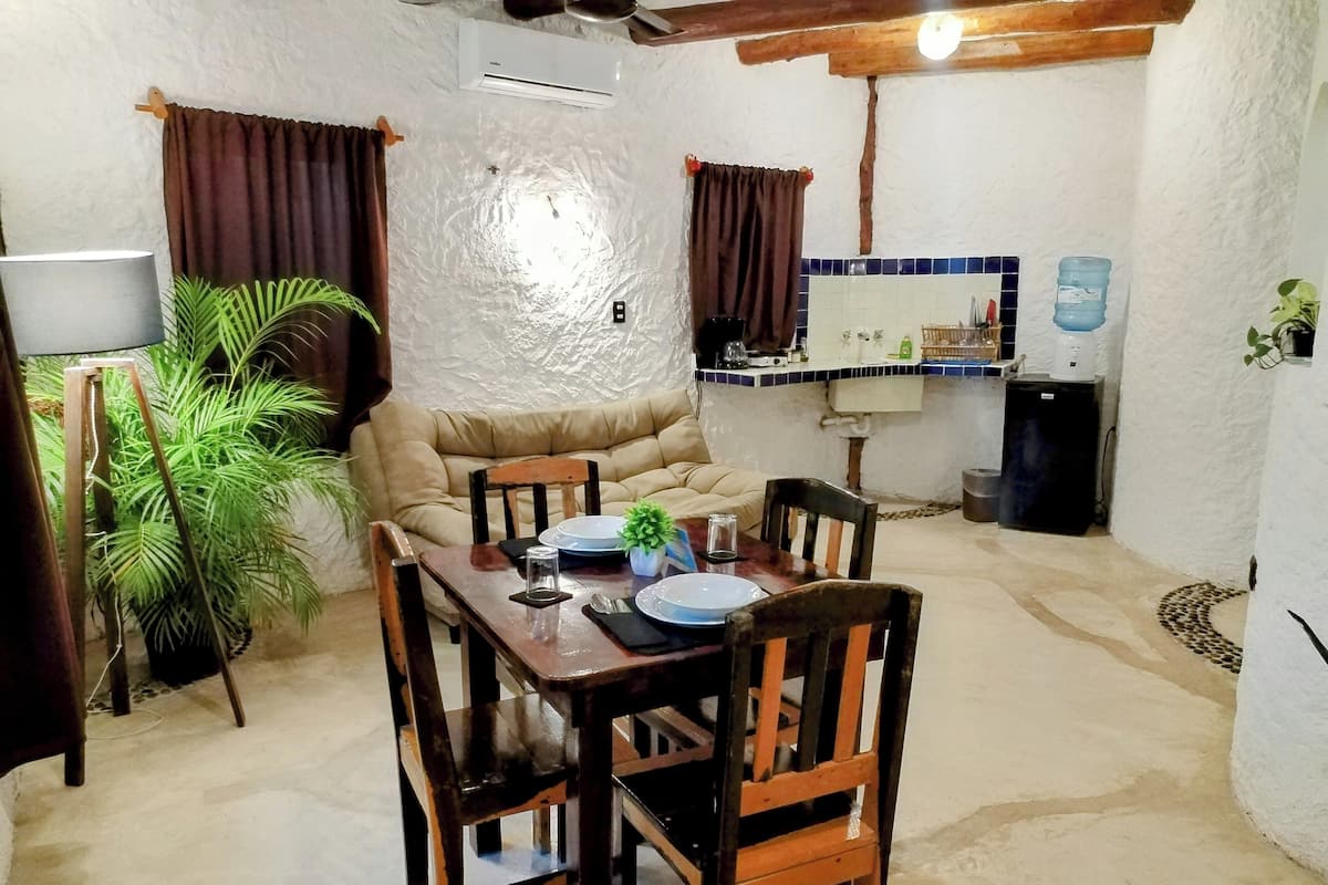 Un coin repas avec une table en bois dressée pour quatre personnes, un canapé et un espace cuisine équipé d’un évier et d’un distributeur d’eau.