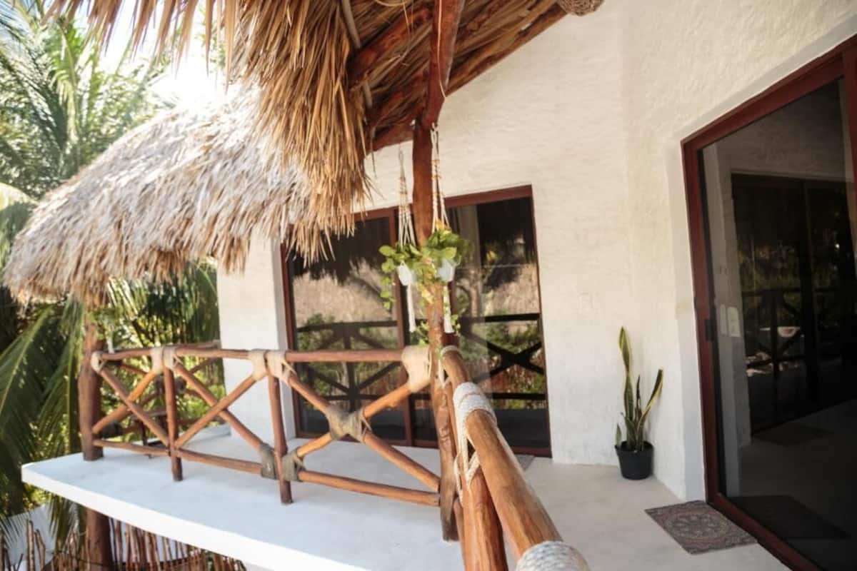 Un espace extérieur doté d’un toit de chaume, d’une terrasse en bois, de plantes en pot et d’une porte vitrée donnant sur l’intérieur.