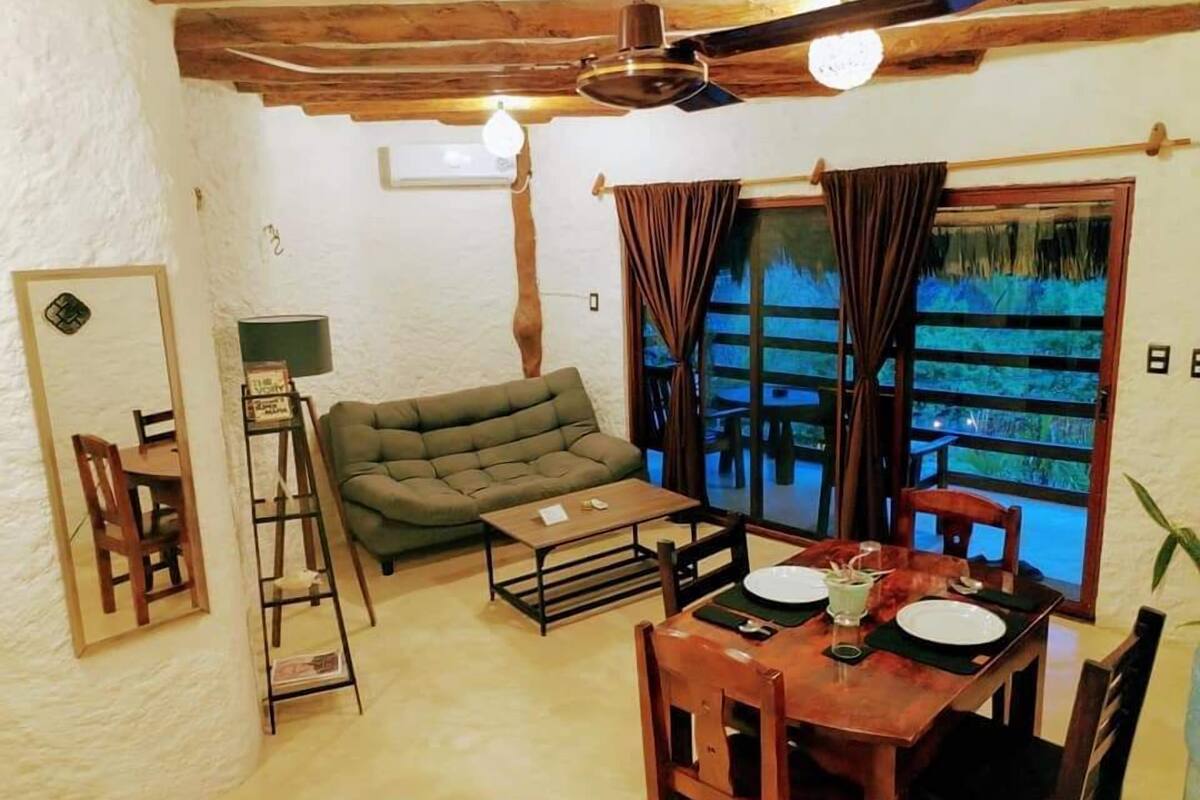 Un salon chaleureux avec un canapé, une table à manger et des chaises, un ventilateur de plafond et une grande fenêtre avec des rideaux.