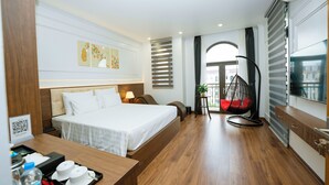 A modern hotel room with a large bed, a desk with a tray of amenities, a hanging chair, and a view of the city through the window. - Azoma Hotel (Hanoi)