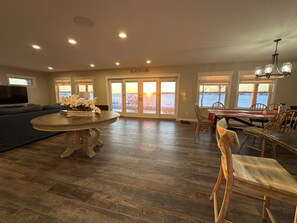 A spacious living room with a large sofa, a dining area with a table and chairs, and a view of a snowy landscape through the glass doors. - Beautiful 4-Bedroom Lakefront Home: dock & hot tub, on White Lake, Whitehall, MI (Whitehall)