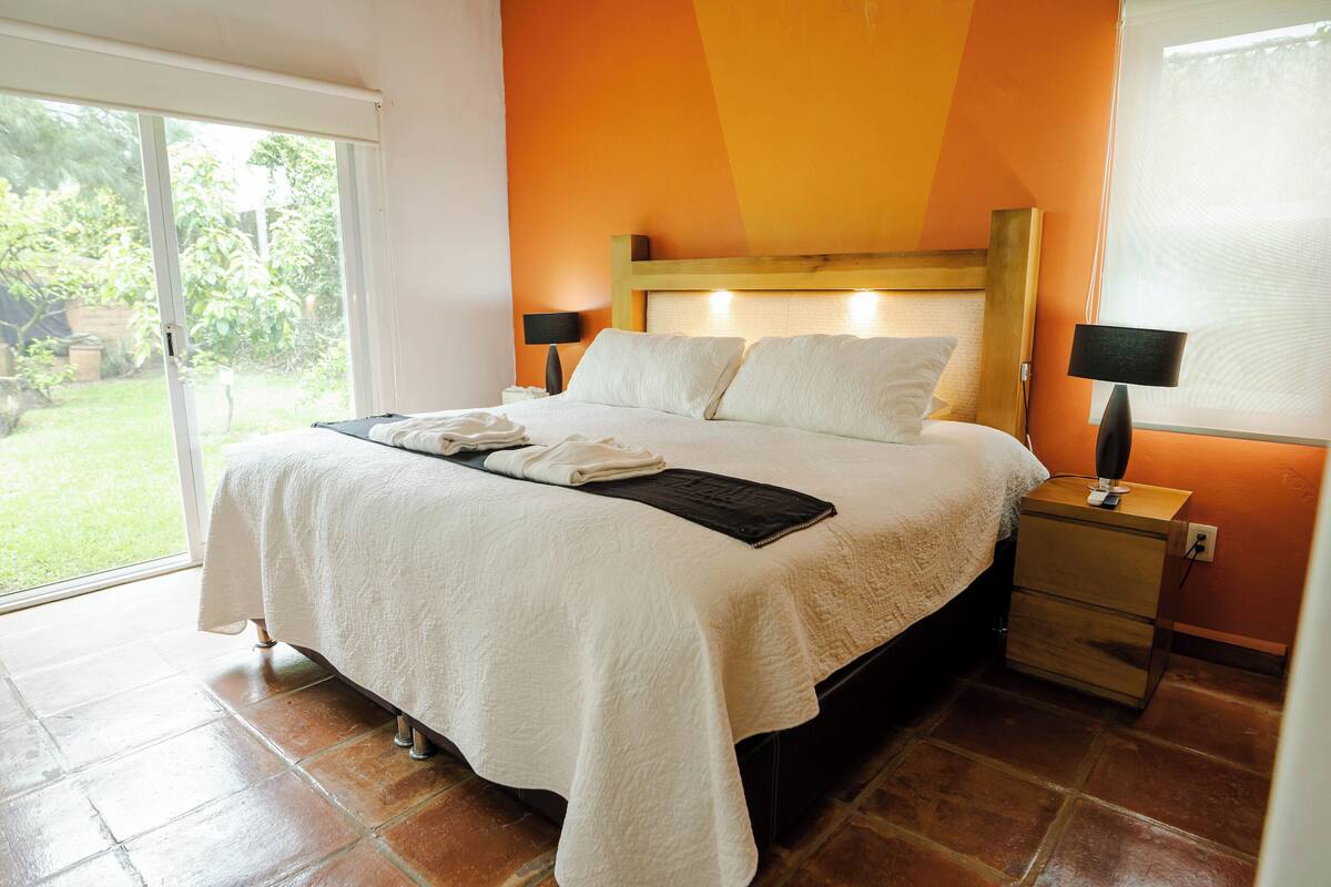 A bedroom with a large bed, two bedside tables, a nightstand, and a sliding glass door to a garden.