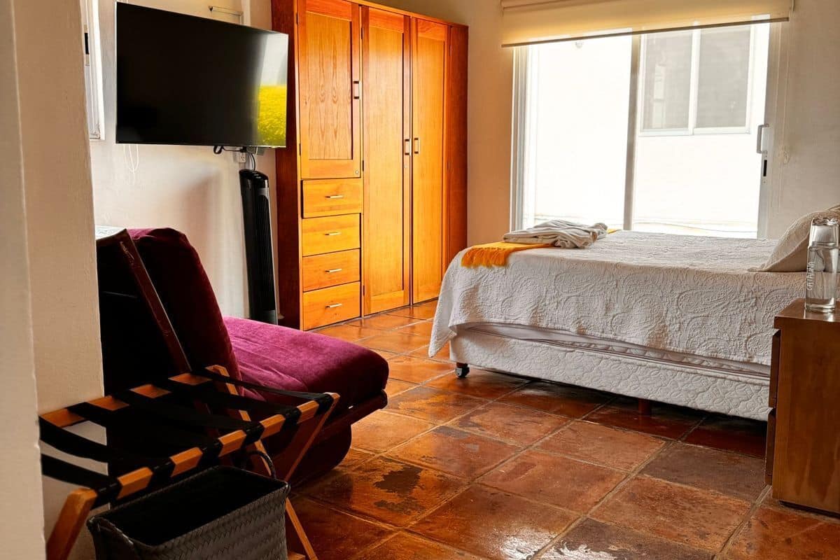 A bedroom with a bed, a wooden wardrobe, a television, and a window with blinds.
