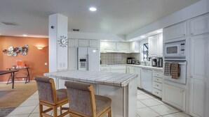 A modern kitchen with white cabinets, a central island, two bar stools, and a dining table with chairs. - Swim & Golf: Palm Desert Home in Senior Community! (Palm Desert)