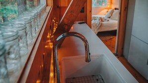 L’intérieur d’un chalet en bois comprend un coin cuisine, avec un évier et une fenêtre sur laquelle sont posés des bocaux.