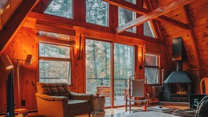 L’intérieur chaleureux d’un chalet, avec un plafond haut, des poutres en bois, une cheminée et de grandes fenêtres offrant une vue sur la forêt.