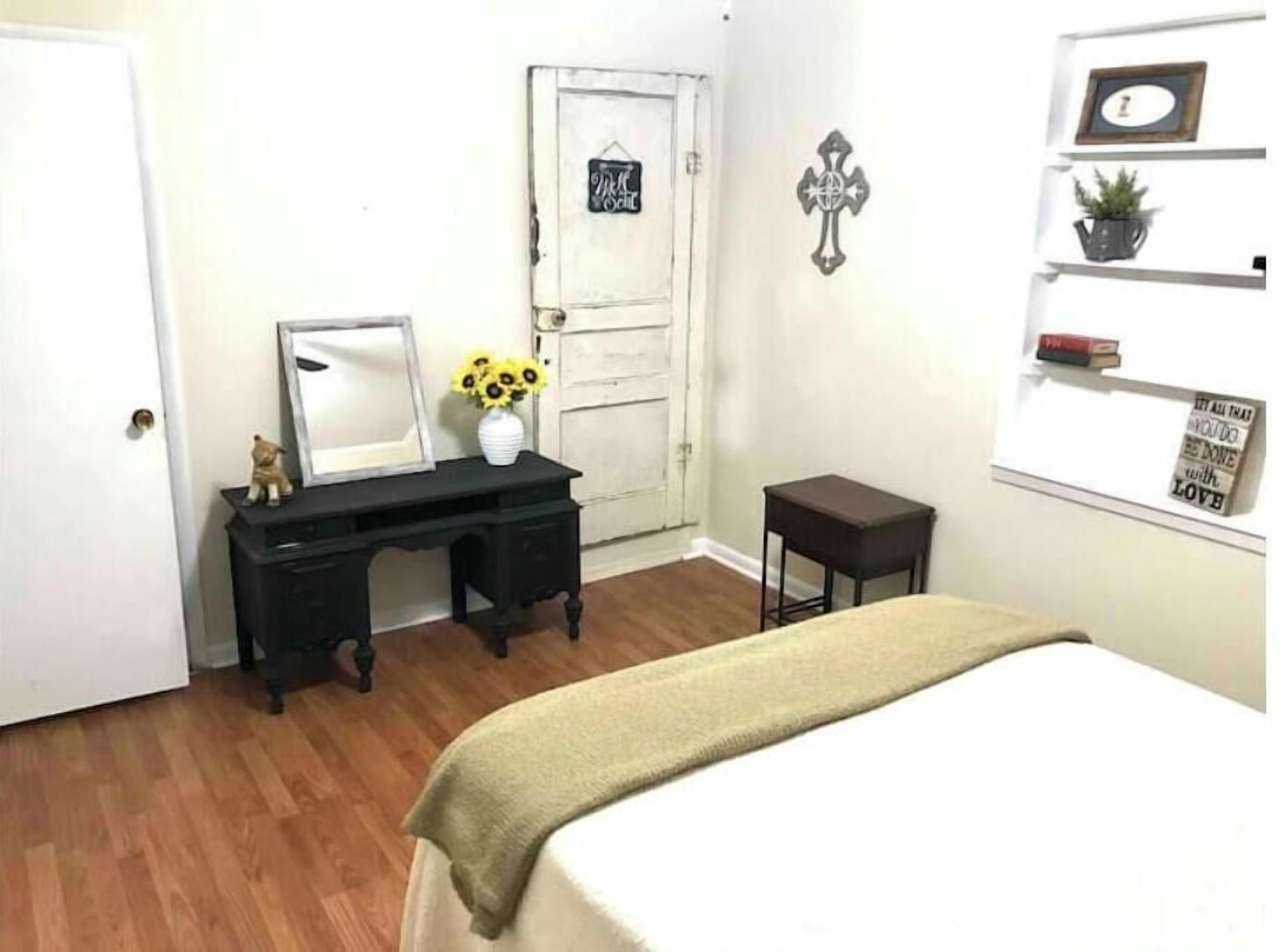 A bedroom with a bed, a desk with a mirror, a small stool, a door, and a wall with a cross and a shelf with a plant and a motivational quote.