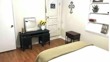 A bedroom with a bed, a desk with a mirror, a small stool, a door, and a wall with a cross and a shelf with a plant and a motivational quote.
