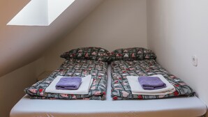 Una habitación pequeña y acogedora, con techo inclinado y tragaluces. Hay dos camas con ropa de cama y almohadas estampadas, y un banco blanco en el centro.