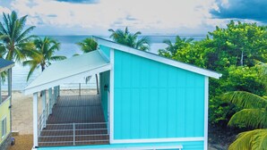 Une maison de plage turquoise à deux étages, dotée d’une terrasse en bois et offrant une vue sur l’océan.