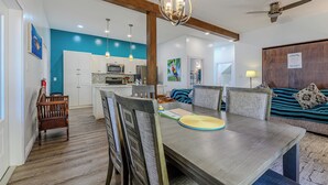 Un coin repas avec une table en bois, des chaises et un banc. Au fond, on aperçoit une cuisine avec des meubles blancs et un mur d’accent bleu.