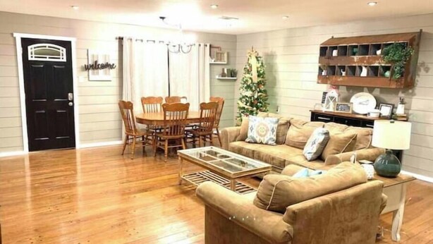 A living room with a wooden floor, a beige sofa, a glass coffee table, a dining table with chairs, a Christmas tree, and a wall-mounted shelf with decorative items.
