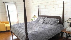 A four-poster bed with a grey quilt, a yellow armchair, and a small table with a lamp.
