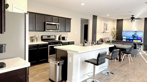 A modern kitchen with dark cabinets, a stainless steel microwave, and a built-in oven. There is a small island with bar stools, a TV mounted on the wall, and a ceiling fan. - Modern 3br/2ba with 3 king beds · Pool · Pets OK! (Patton Village)