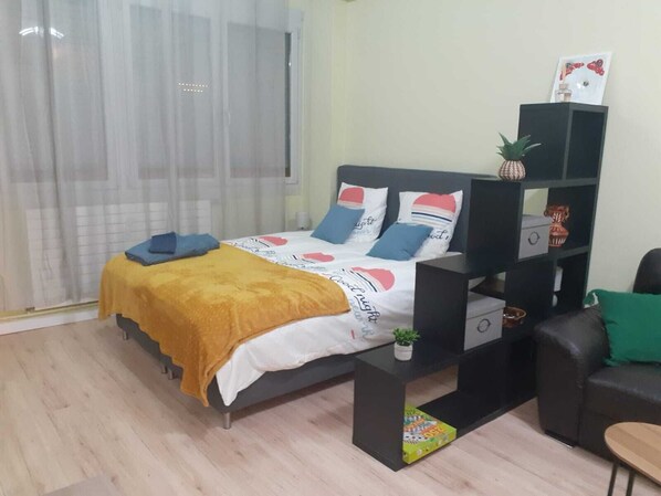 A bedroom with a bed, a nightstand, a small table, a sofa, a window with curtains, and a shelf with decorative items.