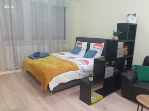 A bedroom with a bed, a nightstand, a small table, a sofa, a window with curtains, and a shelf with decorative items.