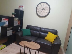 A living room with a black leather sofa, a small wooden table, and a wall clock.