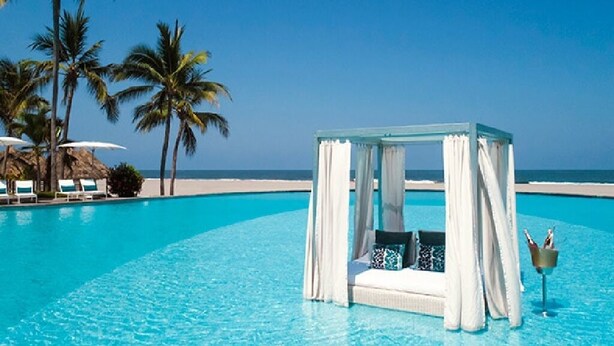 A clear blue pool with a white canopy and a small table with bottles, surrounded by palm trees and lounge chairs.