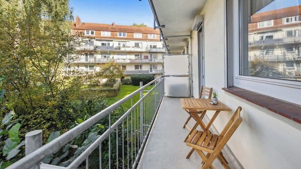 A balcony with a table and chairs, a view of a courtyard, and multi-story buildings in the background.