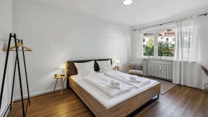 A hotel room with a wooden bed, white linens, a radiator, a window with curtains, and a wooden hanger rack.