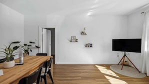 A modern living room with a wooden dining table, black chairs, a flat-screen TV on a tripod stand, and a large window allowing natural light.