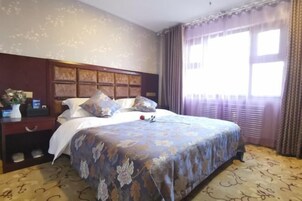 Una habitación de hotel con una cama grande, un cabecero de madera, una mesita de noche con un jarrón y un reloj, y una ventana con cortinas.