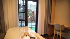 Ein Zimmer mit einem Holztisch, einem Stuhl, einer Tür mit Blick auf einen Garten und einem Fenster mit Vorhängen.
