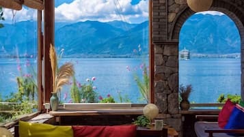 Una stanza con una grande finestra che si affaccia su un lago e montagne, una panca di legno e un tavolo con un vaso.