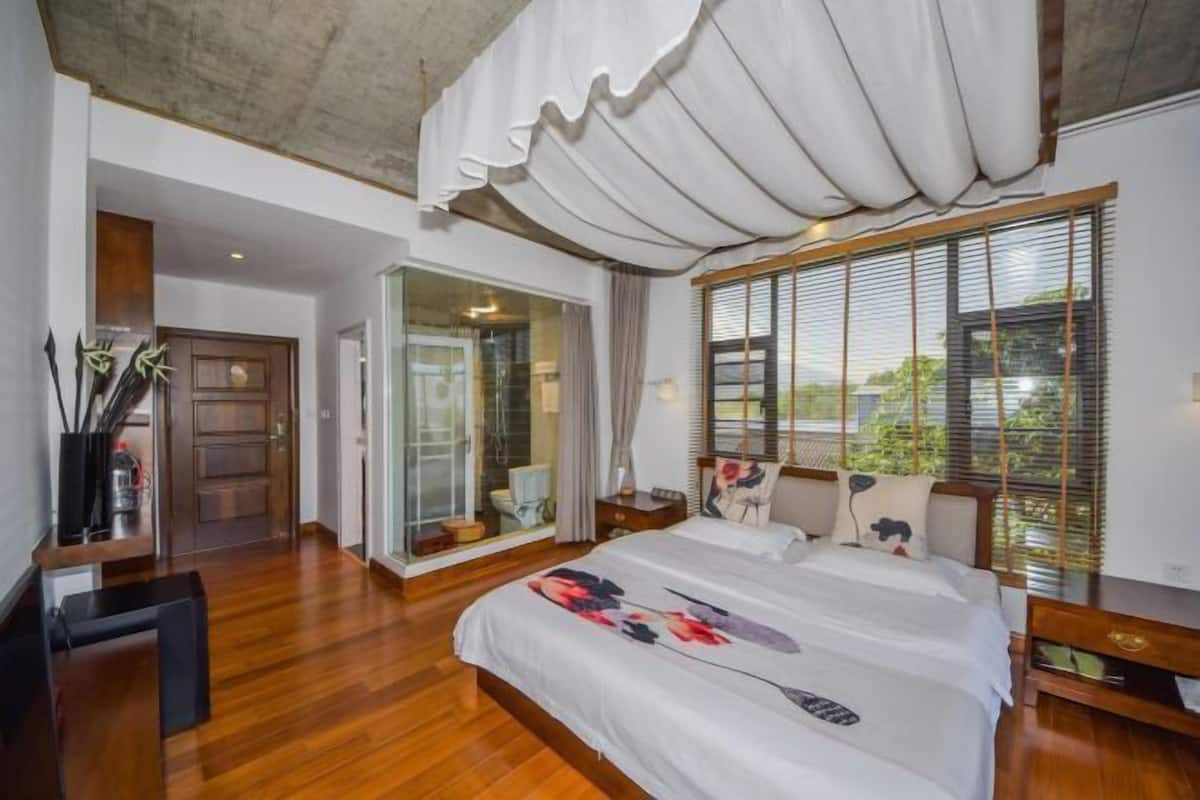 A bedroom with a large bed, a sliding glass door leading to a bathroom, and a view of greenery outside.
