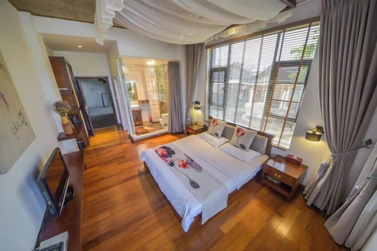 A hotel room with a large bed, a wooden floor, a desk with a chair, a mirror, and a window with curtains.
