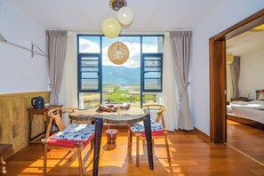 Ein Zimmer mit Holzboden, einem runden Tisch mit Stühlen, Blick durch ein großes Fenster und einem Bett im angrenzenden Raum.