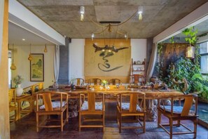 Un espace repas avec une longue table et des chaises en bois, une grande peinture abstraite au mur et une vue sur la verdure à l’extérieur.