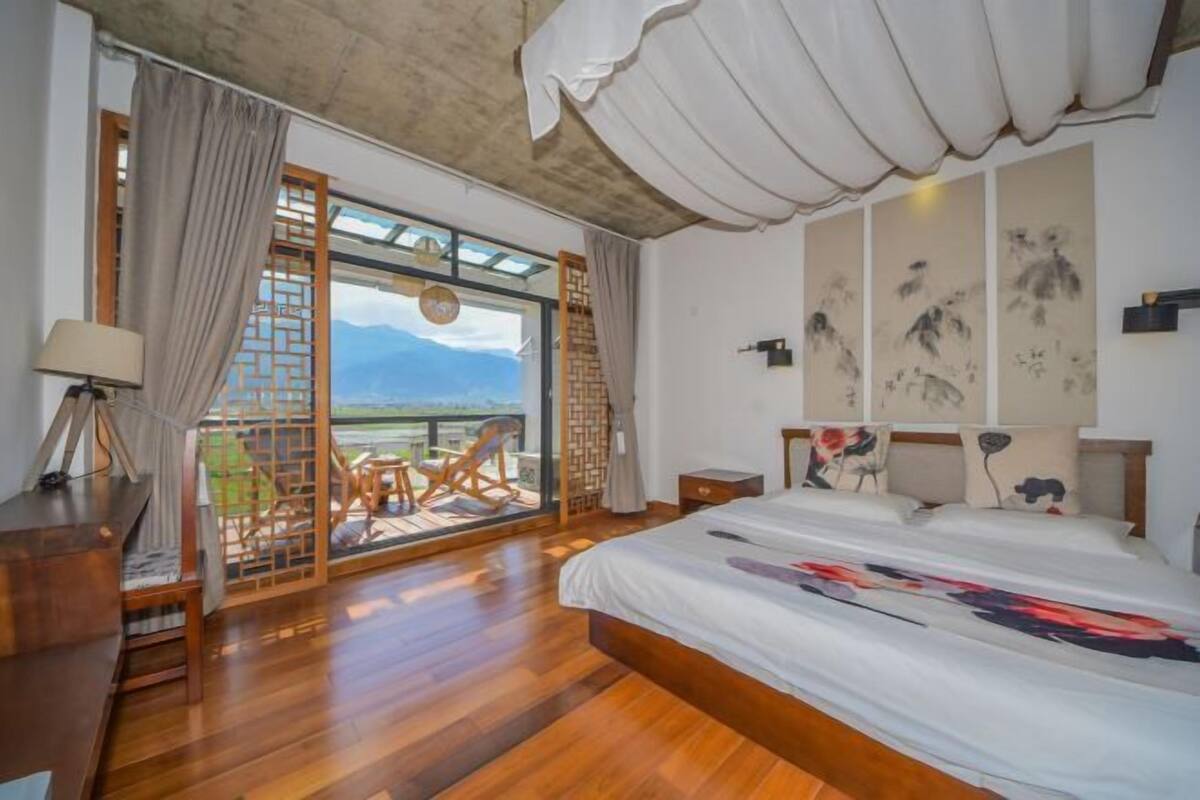A bedroom with a large bed, wooden flooring, a desk with a lamp, a view through a sliding door, and artwork on the wall.