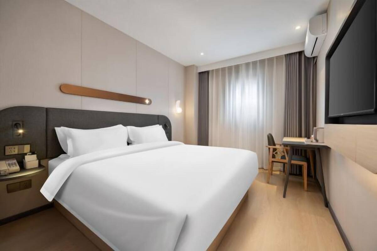 Ein modernes Hotelzimmer mit einem großen Bett, einem Nachttisch mit Lampe, einem Schreibtisch mit Stuhl und einem großen Fenster mit Vorhängen.