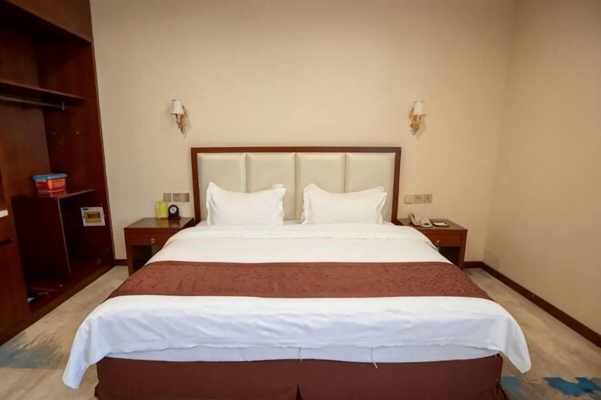 A hotel room with a large bed, two bedside tables, a wardrobe, and a wall-mounted lamp.