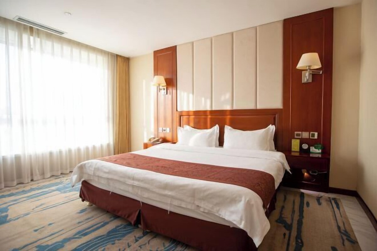 A hotel room with a large bed, a wooden headboard, a nightstand with a lamp, and a window with curtains.
