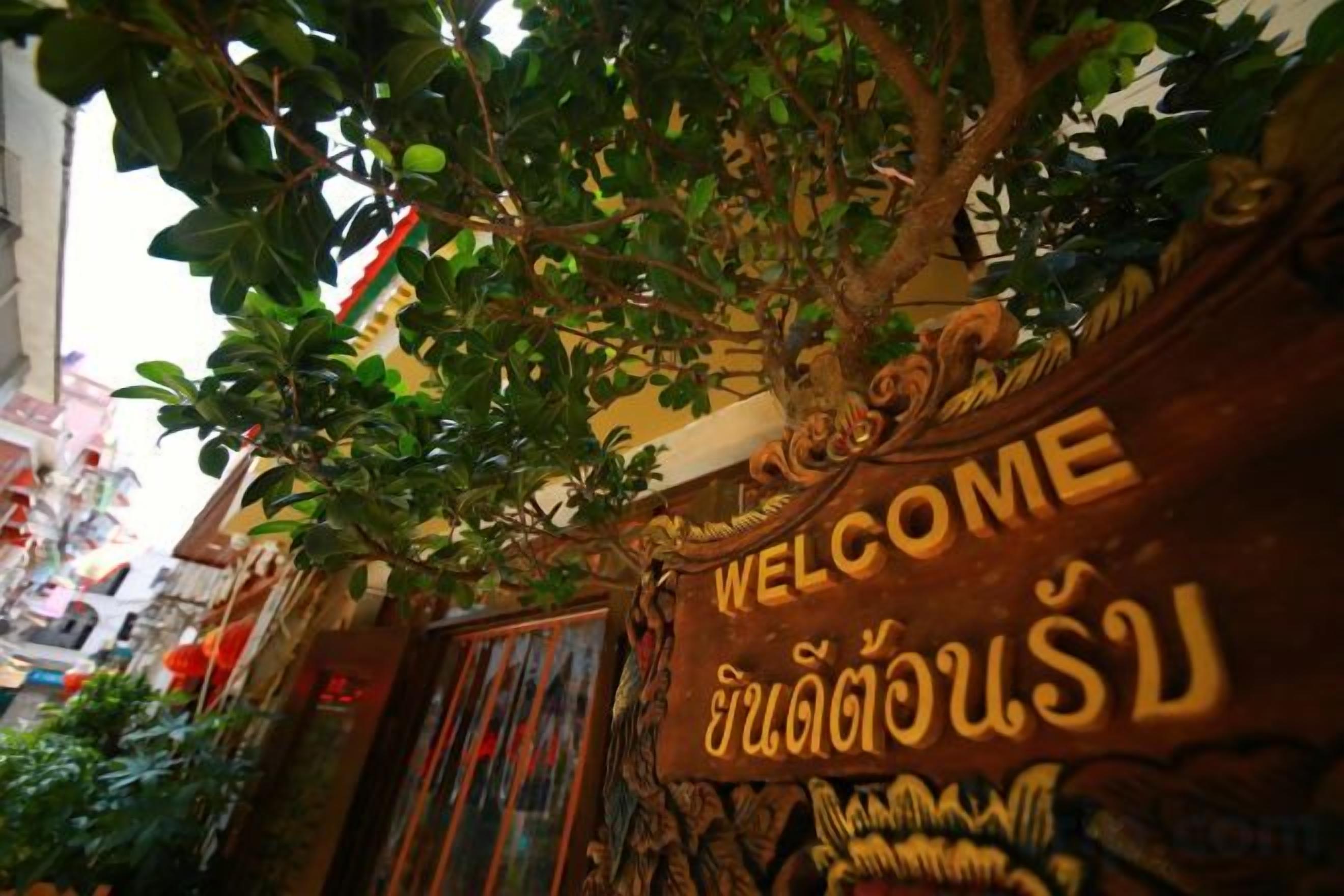 A welcome sign with Thai script, a tree, and a building facade.