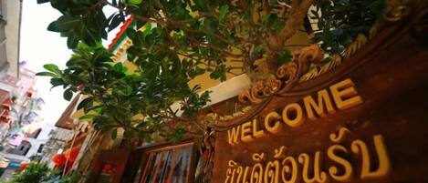 A welcome sign with Thai script, a tree, and a building facade.