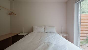 Une chambre à coucher au style minimaliste, avec un grand lit, deux tables de chevet, un banc en bois et une fenêtre donnant sur un paysage verdoyant.