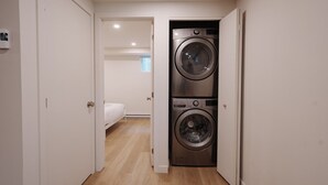 A laundry room with two stacked front-loading washing machines, a bed in the adjacent bedroom, and a white door.