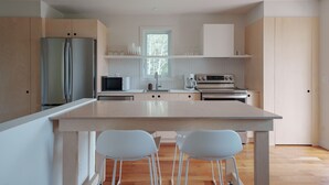 Een moderne keuken met een centraal eiland, twee barkrukken, een koelkast, magnetron, oven, en een raam met uitzicht op bomen.