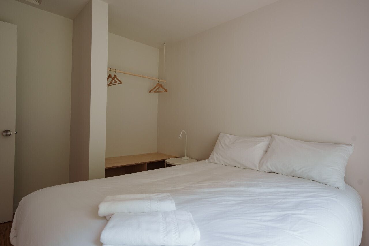 Un letto ben rifatto con lenzuola bianche e due cuscini, un comodino con una lampada e un appendiabiti a parete in legno.