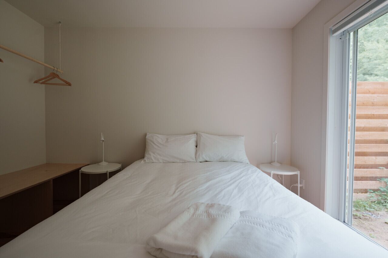 Ein schlicht eingerichtetes Schlafzimmer mit einem großen Bett, zwei Nachttischen, einer Holzbank und einem Fenster mit Blick auf Grünflächen.